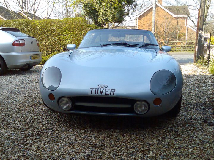 Pictures of your Classic in Action - Page 23 - Classic Cars and Yesterday's Heroes - PistonHeads UK - The image shows a silver sports car parked on a gravel driveway. The vehicle is sleek with a pointed nose and two doors, indicating it's likely a two-seater convertible. It has a visible license plate that reads "TWIGGER." The car appears to be in good condition with shiny bodywork. In the background, there's a partially obscured parked car and a house with a white exterior.