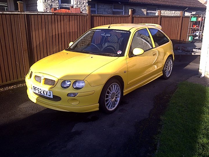 Where is everyone?  - Page 1 - MG - PistonHeads - This image features a large, vibrant yellow hatchback car parked on a driveway. The car is well-lit, likely due to natural sunlight, and its sleek design makes it stand out. It has a curved wheelbase and a sporty flared rear. The driveway appears to be a residential area with a fence and a portion of a house visible in the background. The license plate of the vehicle has a UK format. There is also a person in the background, probably a resident of the home.