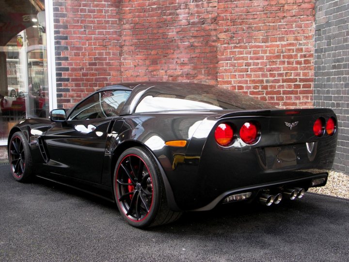 Corvette Z06 Centennial Edition for sale in the UK - Page 1 - Corvettes - PistonHeads - The image captures a scene of a black sports car parked in front of a brick building. The car is poised on a black street, facing the building. Its front is adorned with red detailing and features a low-profile design accentuated by black wheels with red inserts. The building, constructed with red bricks, provides a contrasting backdrop for the sleek black car.