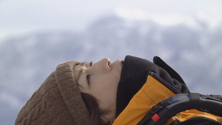 The image portrays a young individual with a contemplative expression. They are dressed in a winter outfit, featuring a beanie and a jacket with a patch, suggesting a recreational or outdoor environment. The person is standing against a backdrop of a blue, possibly cloudy sky, with mountains visible in the distance, giving a serene and majestic feeling to the scene.