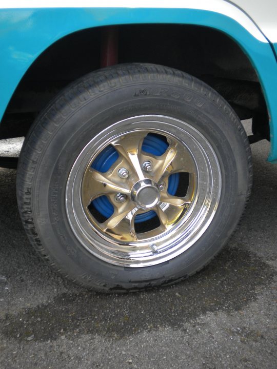 Pistonheads - The image shows a single wheel of a vehicle, prominently featuring a blue and yellow hubcap. This is the sole wheel visible in the frame, captured from a close-up perspective. The background is dark and out of focus, but there appears to be a hint of another vehicle or object partially visible on the left side of the image. There's no text present within this photograph.