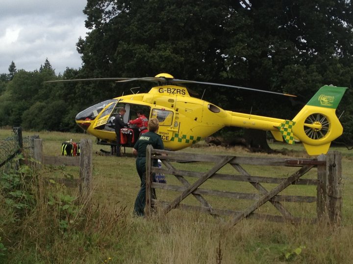 Bagshot Village. Big Incident? - Page 1 - Thames Valley & Surrey - PistonHeads - The image shows a scene with a yellow helicopter on the grass. Through the helicopter's doors, several people are visible, likely the crew. A woman is standing at a gate, which is open, apparently interacting with the helicopter. The surroundings suggest a rural area with trees in the background. There are also some items placed on the grass, indicating the helicopter may have landed there.