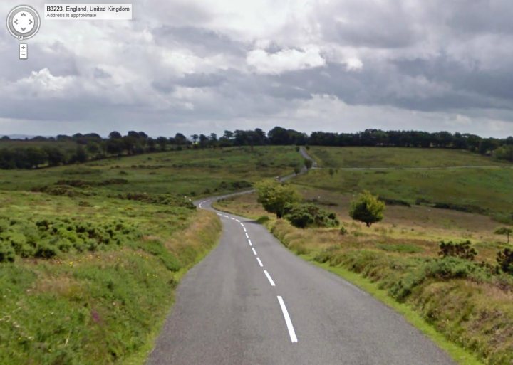 Best bits of roads around Exmoor - Page 1 - South West - PistonHeads - The image is a photograph of a winding road with white dash markings. The road, appearing to be in a rural area, begins in the foreground at the bottom of the image and curves upwards and to the left. It traverses verdant grasslands, dotted with trees and shrubs. The sky above is overcast with dark clouds, suggesting an overcast or possibly gloomy weather condition. In the upper left corner of the image, there appears to be a digital overlay which resembles a GPS marker, indicating the position of the vehicle or person from which the photo was taken, as it reads "A647 England, United Kingdom."