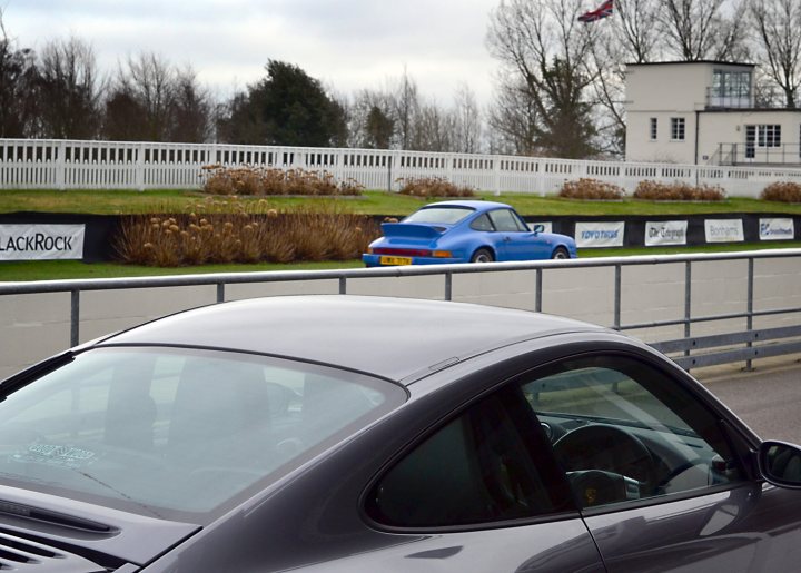 Robert Barrie Track day 24th Feb - photos - Page 1 - Goodwood Events - PistonHeads - The image showcases a dark gray car parked in a lot with white fencing and well-manicured lawns. In the background, there's a track with a blue race car, indicating a racing or auto-show event. Adjacent to the track, a white building with a blue top stands out, which might be related to the event. The scene encapsulates a day at a racing track, possibly a practice day or an event day.