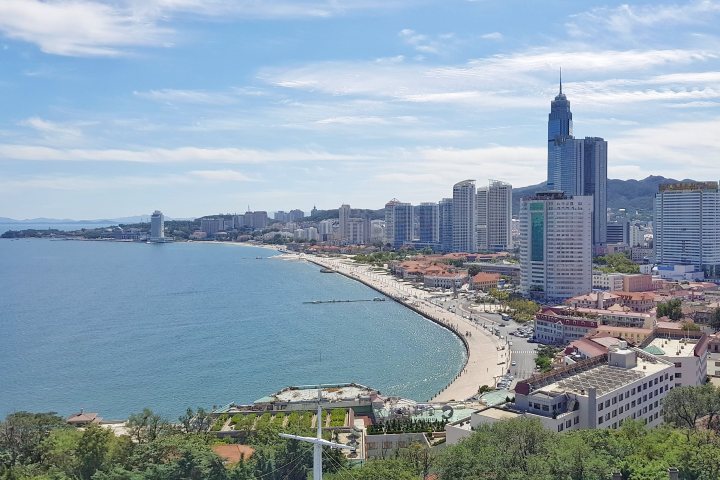 Locations to retire to and why? - Page 11 - Homes, Gardens and DIY - PistonHeads UK - The image shows a coastal city skyline with the ocean and buildings in the background. In the foreground, there's a clear view of a beach with buildings alongside it. There are green trees on the land near the sea, and a boardwalk can be seen. A white building with a balcony overlooking the water is noticeable. The sky above is mostly clear. The image seems to have been taken during daylight, providing good visibility of the cityscape and the ocean beyond.