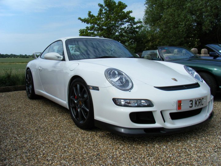 Breakfast Meeting, The Hare, Roxwell 4th June - Page 1 - Kent & Essex - PistonHeads - The image is a photograph of a white Porsche Cayman S parked on a gravel surface. The car is angled towards the left side of the image, showcasing its profile. It features black wheels, sleek bodywork, and a distinctive badge on the trunk. There is a glimpse of a green car in the background and what appears to be a clear sky above. The image has an atypical dark-like tint around the car, perhaps indicating it has been professionally photographed.