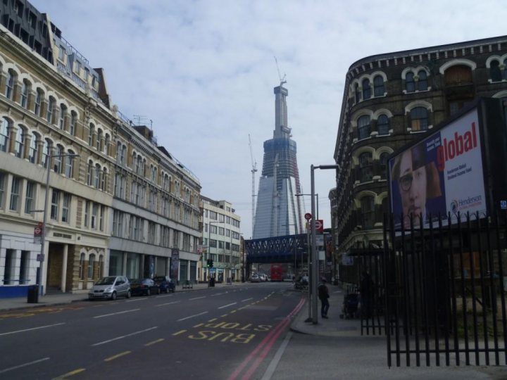 London Pistonheads Tower Tall Built Eiffel - The image captures a bustling European city street, with a mix of old and modern architecture. On the right, two prominent billboards stand against the backdrop of the street, showcasing a woman's face. The architecture to the left, including a church and a large, cylindrical tower, adds a historical charm. The scene is further enlivened by the presence of a few pedestrians and cars, giving it a lively, dynamic feel. A traffic light can also be seen, indicating vehicular traffic in the city.