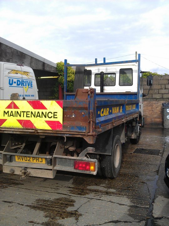 MPG of a fully loaded Artic lorry? - Page 3 - Commercial Break - PistonHeads - The image captures a blue and white commercial truck parked in what appears to be a storage area or alley. The truck is facing the right side of the photo and is equipped with a flatbed trailer. It has a warning sign on its back that reads "MAINTENANCE" with additional text that suggests it's a vehicle maintenance repair service vehicle. In the background, there are other vehicles, including a white van and a brick wall with a pipe running along it.