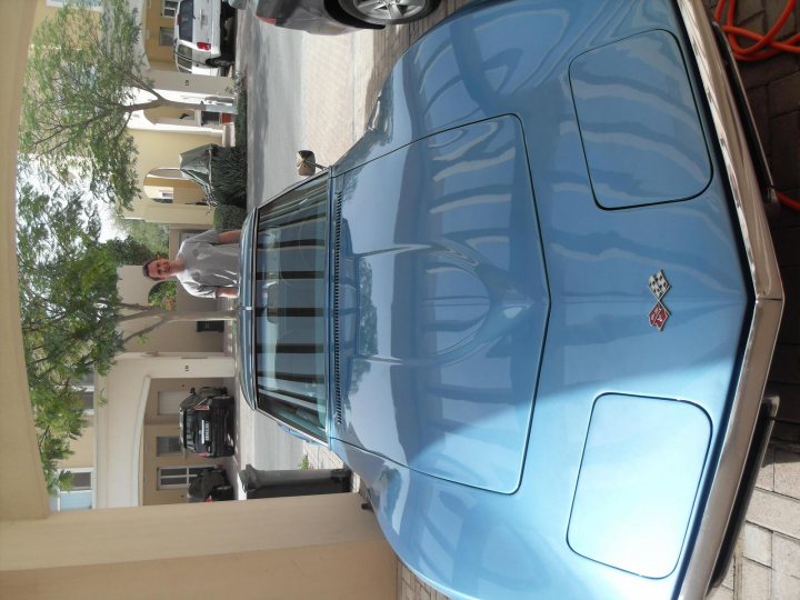 Pistonheads Toy Expat - In the heart of a bustling neighborhood, a man stands off to the side, his figure partially obscured by the blue sports car that takes center stage in this image. The car, sleek and shiny, is parked under the shelter of a covered patio, its hood gleaming. Adjacent to the patio, a brick wall adds texture to the scene, while a tree stands tall in the background, providing a touch of nature amidst the urban setting. In the distance, a few other cars are parked, hinting at the daily hustle and bustle of this neighborhood.