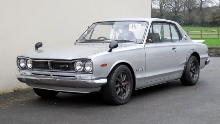 Hakosuka GTR - Page 1 - General Gassing - PistonHeads - This image shows a medium-sized, compact car with an open hood parked on an asphalt surface next to a building with a white exterior. The car appears to be a vintage model, as suggested by the style of the vehicle, including the distinctive headlights, square, pillarless body design, and the large emblem on the front grille, which is possibly a logo of a well-known automaker. The vehicle is equipped with five-spoke wheels that add to its vintage appeal. The image is color, with no visible texts or branding that provide further context about the car or its location.