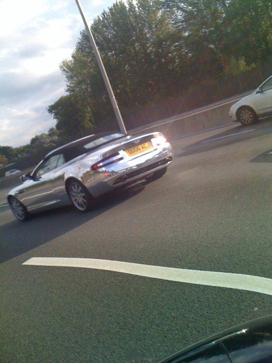 Pistonheads - This image depicts a scene from a highway. The focus is on a metallic silver car driving away from the viewer. The car is adorned with a yellow license plate that reads "B81 MH". The car is distinguished by its white plates with a black font. The highway is lined with trees, and there is another car visible in the distance, slightly ahead of the silver car, adding depth to the scene.