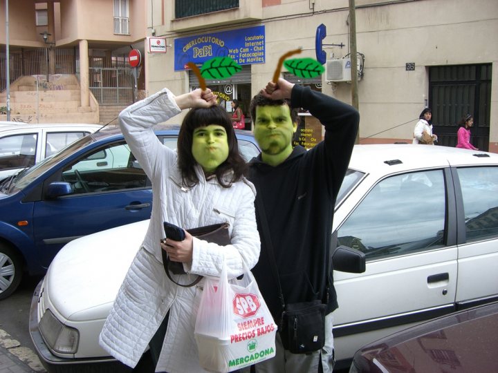 The image is a humorous and quirky street scene. Two people are the main subjects; one is dressed in a white jacket, wearing a green mask with exaggerated features that make them look like they have a large, flapping mouth with a wide smile. The other is in a black hoodie, wearing a mask that closely resembles the green one, suggesting that they might be part of the same group or event. Both are holding up green masks towards the camera, giving a playful impression. They are positioned in front of a blue car and a building with a sign that appears to be in a foreign language, and there are other cars and people in the background. The overall ambiance suggests a lively, possibly urban environment.