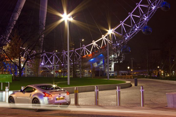 Photos Session In London - Page 1 - Photography & Video - PistonHeads - The image captures the lively night-time atmosphere of a city. A sleek gray Mazda MX-5 Miata can be seen parked on the side of a curb. The street is illuminated by bright streetlights and a neon sign. In the background stands the iconic London Eye, a massive Ferris wheel, which glows with lights, adding to the festive night scene. The street appears to be in a bustling city area, brimming with life.