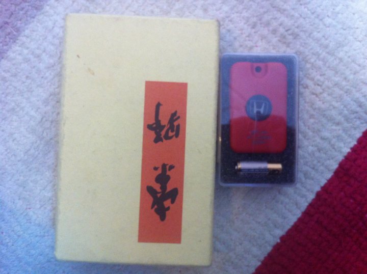 What shall I do with this? - Page 1 - Honda - PistonHeads - The image shows a small red smartphone lying on the ground beside a larger cardboard box and a clear plastic tablet case. On the top of the box is a rectangular orange and black label, possibly containing Japanese text, suggesting that the items could be of East Asian origin. A thin vertical line runs parallel to the phone and the label on the box in both cases. The phone is resting beside the paper towel or napkin on the floor, indicating the functioning of the phone in this grassy area. The overall scene conveys a casual and uncluttered environment, possibly as part of outdoor or travel activities.