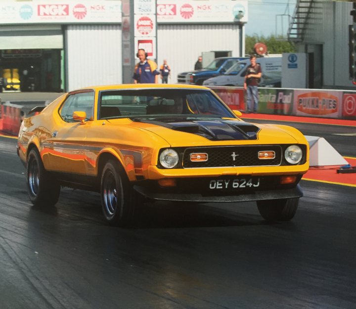 My Mach 1 showing a souped up Fiesta some dust - Page 1 - Mustangs - PistonHeads - The image shows a vibrant scene at a car show or race track. A bright yellow Mustang muscle car, possibly from the late 60s or early 70s, is the focal point of the image. It's on a track or a street with safety hazards like warning and caution signs visible in the background. Two individuals appear to be inspecting the car, their attention captured by its detailed and shiny features. The setting suggests an enthusiastic atmosphere filled with vintage vehicles enjoying the public's admiration.