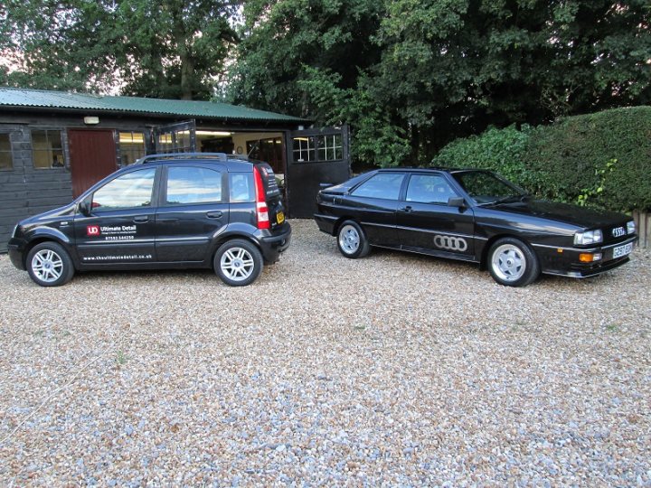 A car parked in a parking lot next to a car - Pistonheads - The image showcases a set of wheels and a car, which have been carefully aligned and photographed in a clear, outdoor setting. The car is a classic 90s, black, four-door hatchback with other features like a spoiler and a low license plate. It appears to be in excellent condition, suggesting that it is well-maintained or has been recently restored. 

Standing next to the car, there's another vehicle of the same model and same body color, contrasting with its white-rubber wheel and silver trim. The parking lot is made of gravel or fine gravel, indicating it might be a quiet, less-frequented area. The setting seems to be a private or semi-private space, likely associated with the car owners, given the calm and organized representation of both the wheels and the car.