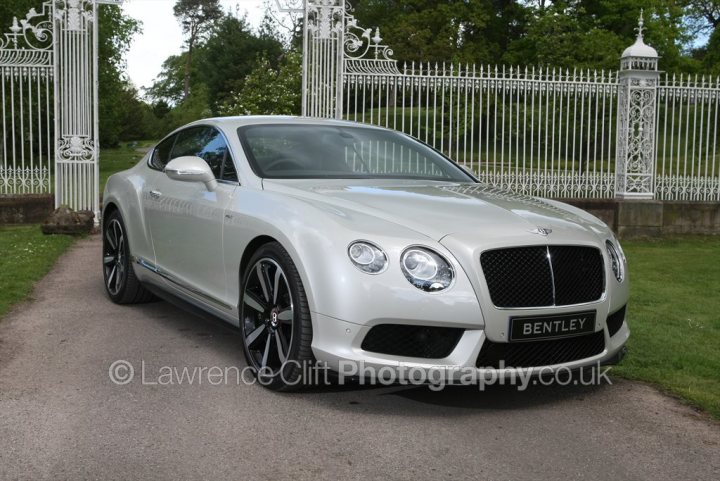 Cholmondeley Pageant of Power 13-15 June - Page 1 - North West - PistonHeads - The image presents a sleek, silver Bentley car parked in front of a large, wrought-iron gate. The gate's intricate detailing provides a striking contrast to the car. On top of the gate, there is a sign with some text, which seems to be associated with the venue or location. The car is situated on what appears to be a gravel or dirt road, possibly leading into a driveway or parking area. The scene is well-lit, suggesting a clear day with no weather issues. The overall impression is one of elegance and privacy.