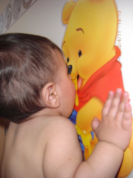 The image captures a tender moment between a small child and a character from the Winnie-the-Pooh series. A baby, who appears to be very young, is lying face down on a yellow surface with a paper later placed over it. The baby's pudgy cheeks and sweet expression are the focus of the image. In this playful interaction, the baby is pressing his face against the cartoon cutout of a popular character, seemingly trying to give the character a kiss. The scene evokes a sense of delight and innocence, as the child innocently engages with a beloved character. The image is a snapshot of childhood curiosity and the simple joys that come from discovering new things.