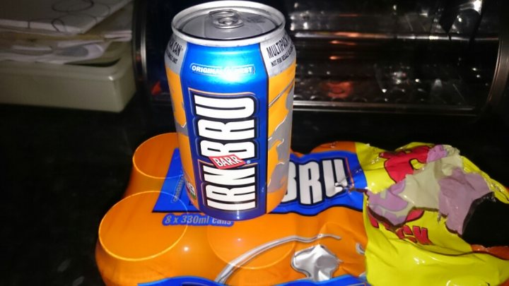 A hot dog and a drink on a table - Pistonheads - The image captures a casual kitchen scene with a can of Irn-Bru, a popular Scottish soft drink, as the centerpiece. The can, bearing an iconic blue, orange, and yellow color scheme, is prominently featured in the foreground, evoking a sense of refreshment. Further down, a packet of Walker's Pure Gold deep-fried potato chips and an unidentified snack are also visible, suggesting a classic approach to satisfying one's hunger. The kitchen appliances and the detritus of daily life in the background paint a picture of a well-lived-in space, adding a touch of homeliness to the overall scene.