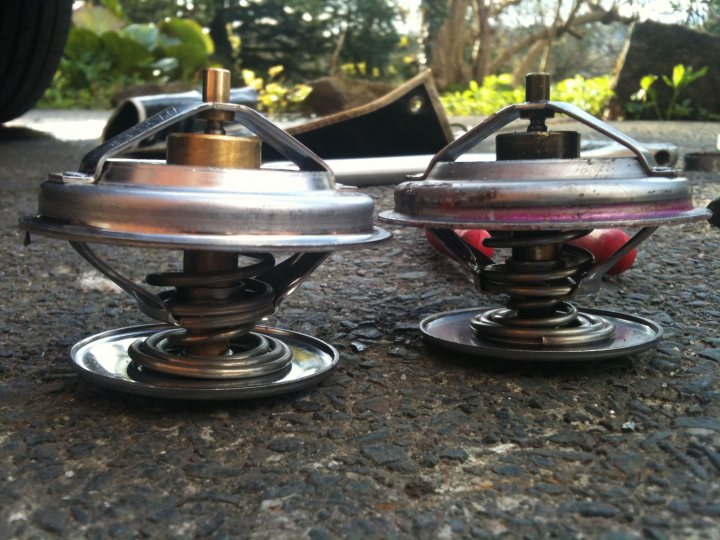 Cleaned Pistonheads Today - This image shows a pair of used car radiator fans side by side on what appears to be an asphalt surface. The fans have metallic parts, with some of these components appearing to be worn or damaged. The centers of each fan feature copper-like padding, and there is visible rust or corrosion in places. In the background, there is a hint of greenery and a metal object that might be part of some machinery or equipment, suggesting that this scene takes place in a garage or an outdoor maintenance space.