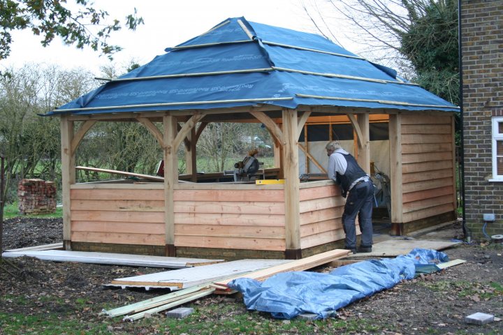 Framed Pistonheads Garage Building Oak Garden - The image shows a small wooden structure with a blue tarp covering its roof. There is a person standing inside the cabin, wearing a white helmet. They are standing next to a table with various tools scattered around. The person appears to be working on the construction or renovation of the structure. The area surrounding the cabin is strewn with construction materials such as lumber and various other tools. The ground is uneven and covered with debris, suggesting ongoing work.