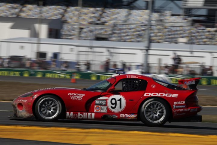 Daytona 24hr tour  - Page 1 - Vipers - PistonHeads - In the center of the image, a vibrant red Dodge Viper V10 RS racing car is prominently displayed, driving on a race track. The car is adorned with a white number 91 prominently displayed on the side, adding to its racing aesthetic. The backdrop of the image reveals a grandstand filled with spectators, suggesting an ongoing racing event. The race track is bordered by green grass, offering a contrasting color to the red vehicle. The perspective of the image suggests it was taken from a camera positioned low, capturing the car in motion from this angle.