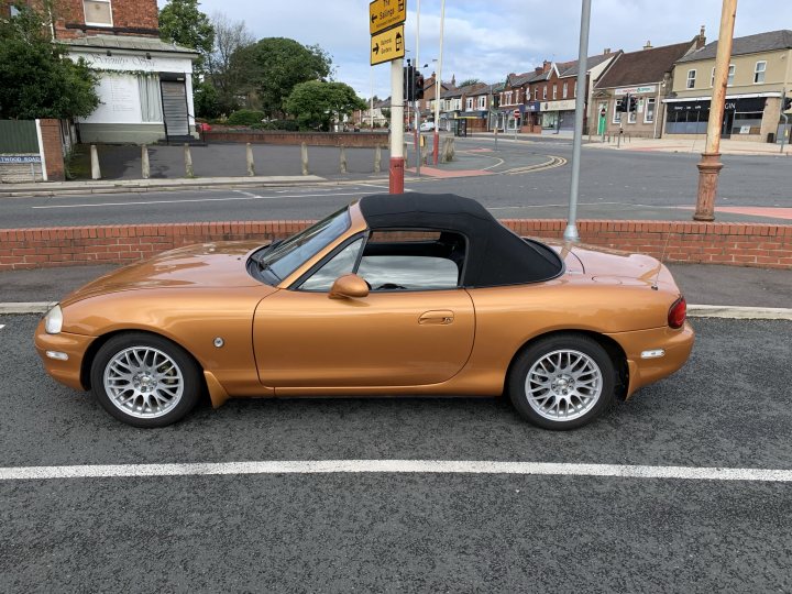 1999 MX5 Restoration  - Page 1 - Mazda MX5/Roadster/Miata - PistonHeads UK - The image shows a vintage convertible car parked in what appears to be a parking lot. It is painted in a shade of gold or tan, with a black soft-top roof and convertible hood. The car has white wheels and a prominent rear spoiler. The vehicle's bodywork looks well-maintained, indicating it may be a classic or special edition model. In the background, there are buildings visible under a partly cloudy sky. The overall style of the image is that of an everyday photograph with no artistic manipulation. There is no visible text in the image to provide additional context.