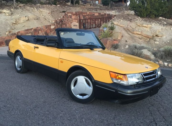 Lovely Cars: Interesting, Classic, Retro, Barge 5-10k - Page 59 - General Gassing - PistonHeads - The image is a photograph of a yellow coupe parked outdoors. The car features a retro design with a curved windshield and a distinctive rear end, indicative of models from the 1980s or early 1990s. It has a black convertible top down, showing two black seats visible from the rear perspective. The body of the car seems well-maintained, with glossy paint and clean lines. The car is stationed on a smooth surface that appears to be a part of a driveway, with a sloped ground and sparse vegetation in the background.