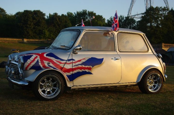 London Brighton Pistonheads - The image captures a vintage Mini Cooper car displayed on a field, likely during a car show or exhibition. The car's body is silver, gleaming under the light, and it is adorned with a striking British flag graphic on the side and hood. The flag features red, white, and blue stripes, a classic symbol of British nationality. A single Union Jack flag is mounted on the hood, and on top of the trunk, there are two flags. The setting sun in the background casts a warm glow over the entire scene, creating a serene and picturesque environment for showcasing the car.