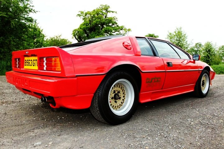 Pistonheads Pick Late - The image shows a red sports car parked on a gravel surface. The car is of a vintage design, suggesting that it is an older model. It has a prominent front bumper and a rear spoiler, typical features found in sports cars. The car's distinctive style, combined with the gravel surface, suggests that the vehicle is in an outdoor or rural setting. There are trees near the car, indicating that it is parked in a natural or semi-rural environment.