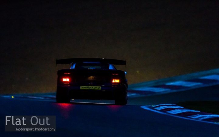 Brands Hatch Timeattack - Page 1 - Noble - PistonHeads - The image shows a nighttime racing scene with a car. The car is on a racetrack, illuminated under its own headlights which are on. The racetrack has white stripes marking the lanes, enhancing the dramatic racing atmosphere. The color palette of the image mainly consists of black and blue with the headlights adding a stark contrast. There is text imagining over the picture, providing additional context or information. The image also has a watermark in the bottom left corner, indicating the source or ownership of the photograph.