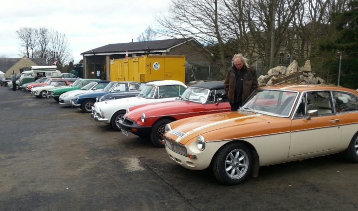 Bellingham classic car show - Page 1 - North East - PistonHeads - The image showcases a row of vintage automobiles, neatly lined up on a black asphalt road. The cars, with their side windows and vintage design, all face the same direction, suggesting a gathering or a show of classic vehicles. The man in the green jacket, positioned in the middle, appears to be observing the parked cars. In the distance, on the right side, there's a yellow bin, adding a touch of color to the scene. The backdrop features trees and a building, providing a natural and urban setting for this collection of classic vehicles.