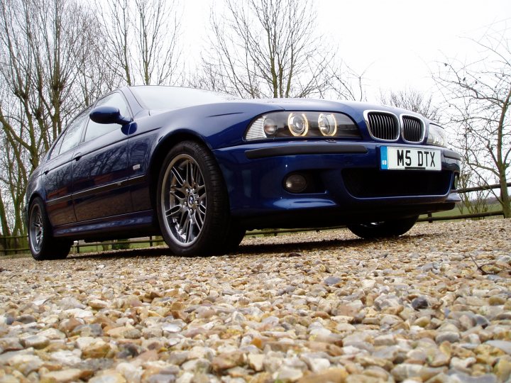 Pistonheads - This image captures a moment on a gravel path where a sleek blue BMW M5 sits parked. The car is positioned facing the camera, showcasing its unique design features. The license plate, reading "M5 DTX", is visible, along with the distinctive headlights and BMW logo. In the backdrop, the earthy tones of the gravel path contrast with the car's metallic blue color. The surrounding leafless trees suggest a late fall or winter setting. The image conveys a sense of tranquility and the timeless beauty of this high-performance vehicle in a serene natural setting.