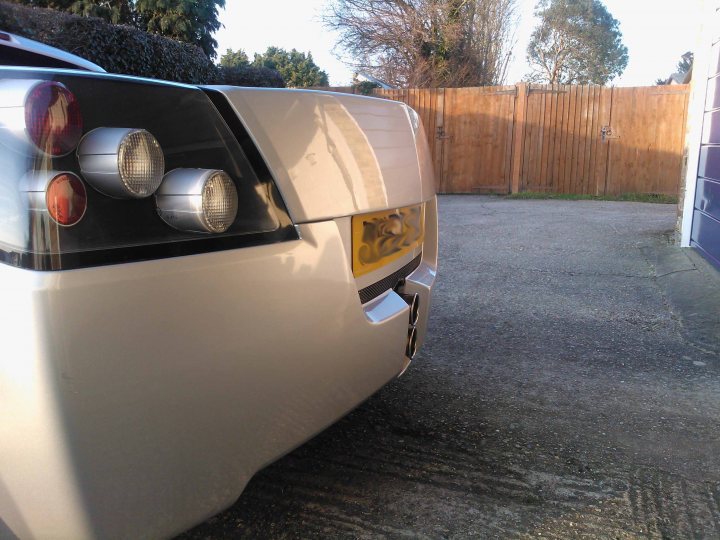 Pistonheads - In the image, a silver coupe is parked on a gray pavement. The coupe is positioned facing the right side of the image, while the sun is partially obscured by a tree to the left, causing it to be cast in shade. The back of the coupe is adorned with a set of red and silver signal lights. The fence in the background complements the overall tranquility of the scene. The relative positions of the car, the sun, and the tree create a vibrant dynamic.