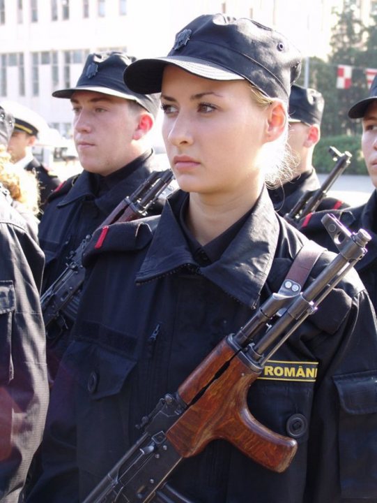 Gun Girl - The image depicts a young person, possibly a woman, donned in a black uniform. She carries a rifle slung over her shoulder, a symbol of her role or occupation. Her expression is serious, suggesting a corresponding sense of responsibility or duty associated with her attire. The background is blurred, drawing focus to the individual in the center.