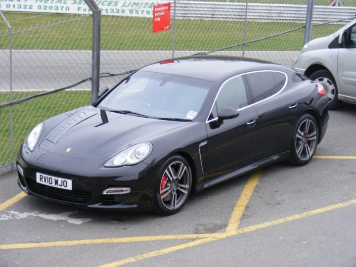 Rarities Pistonheads Spotted Supercars - The image showcases a black Porsche car parked in a lot, with its glossy exterior gleaming under the sunlight. The car is positioned on a yellow parking line, highlighting the contrast between the vehicle and the ground. Interestingly, a red warning sign is prominently placed in the vicinity of the car, and there is an open metal gate in the background. Another car is parked a bit further in the background, indicating the presence of other vehicles in the area.