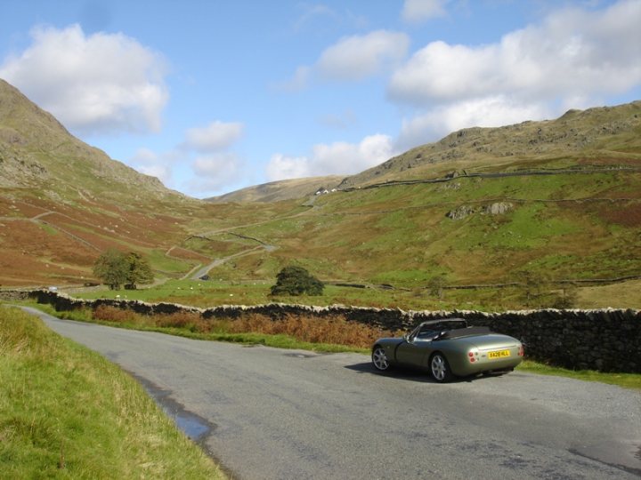 Help identify this Lake District location? - Page 1 - General Gassing - PistonHeads - The image captures a tranquil scene on a country road. A sleek, silver-colored sports car is parked on the gray asphalt road, facing the camera. The road stretches ahead, leading the eye deeper into the landscape. The road is bordered on both sides by a traditional stone wall. Above the road and wall, a valley filled with lush, green hills rolls away, dotted with patches of brown and cattle grazing in the distance. The hills are crisscrossed with winding roads and a few buildings, suggesting a rural or countryside setting. The sky overhead is blue and dotted with white clouds, creating a serene and picturesque backdrop to the scene.