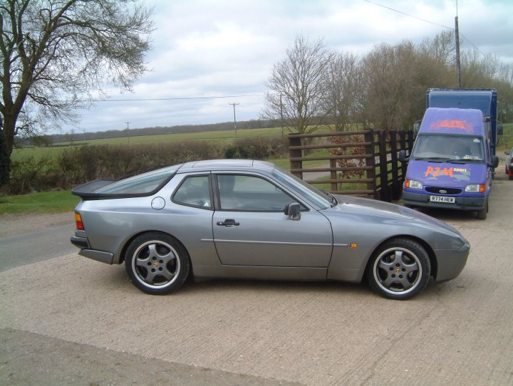 Frontrunner Pistonheads Loves Warning - The image shows an outdoor scene with two vehicles and a person visible. In the foreground, there is a silver-colored sports car with a black rear windshield and a purposeful yet elegant design. The black rubber roof and the prominent red-orange tail light stand out against the vehicle's body color. Behind the car is a robust, black metal gate with a fence structure atop it, followed by a large, open field that extends into the distance.

On the right side of the image, there is a purple van with a white lettering. The van is parked and appears to be facing towards the open field. The vehicle is stationary and is not atop a parking structure. The field itself is expansive and grassy, contrasting with the concrete surface on which the car is parked.

In the top right corner, there is a person wearing a blue jacket, partially obscured by a large gray pole. The person seems to be outside in the daytime and is not directly interacting with either of the vehicles. The presence of the person, along with the open field and gate, suggests this could be a rural or semi-rural setting. The sky above is overcast, giving the scene a cool,