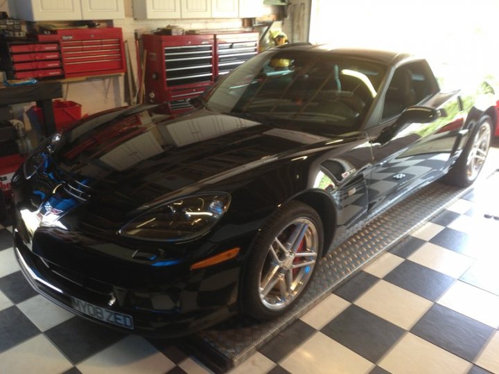 Z06 day today - Page 1 - Corvettes - PistonHeads - The image features a sleek black two-door convertible car parked in a garage. The car's top is down, revealing a shiny black interior. It's sitting on a black lift gate, positioned on black and white checkerboard tiles. In the background, there are several tools and equipment stored in the garage, indicating a well-equipped space for vehicle repair and maintenance.