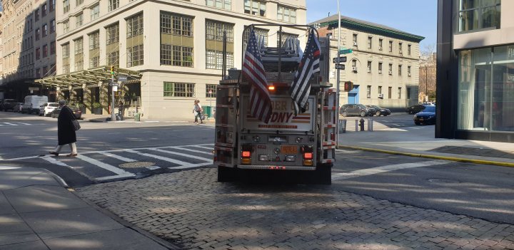 Touristy Film Locales.   - Page 5 - TV, Film & Radio - PistonHeads - The image shows a scene on a city street. A fire truck is prominently featured in the foreground, parked next to what appears to be an intersection. The vehicle has red and white lights on top, indicating that it may be an emergency response unit. Behind the fire truck, there are buildings that line the street, suggesting an urban environment. On the sidewalk, a person can be seen walking, adding life to the scene. In the background, a traffic light is visible, further emphasizing the urban setting. The sky above is clear and blue, indicating good weather conditions at the time the photo was taken.