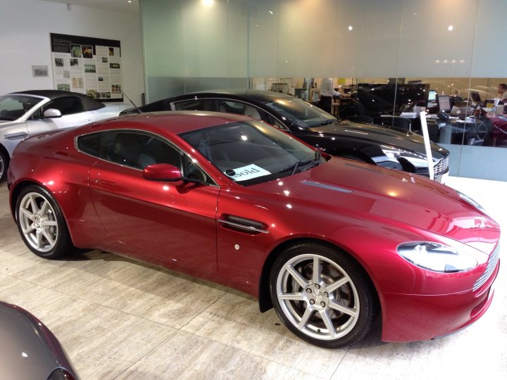 Collecting my first Aston on December 28th!! - Page 5 - Aston Martin - PistonHeads - The image displays a vibrant red sports car prominently in the foreground, reflecting off the smooth marble floor. Behind the car are other cars, suggesting that the vehicle is in a showroom or dealership. The glass window allows a view into the adjacent room where people are visible. The overhead lighting and clean environment indicate a professional setting.