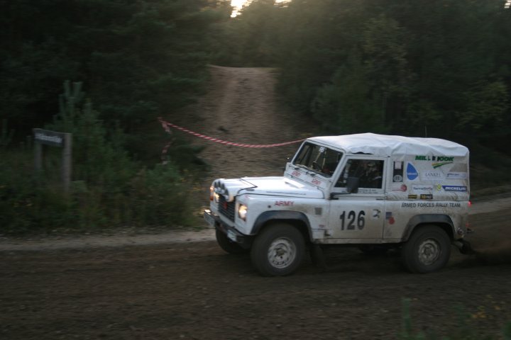 Tempest Rally tomorrow. Anyone on here going? - Page 1 - Thames Valley & Surrey - PistonHeads - The image is a photograph showing an off-road vehicle, specifically a four-wheeled truck, in motion on a dirt track. The vehicle appears to be covered in dust, indicating recent use on the trail. It is equipped with large black tires suitable for off-roading and has various decals or markings on its bodywork. In the background, there's a signpost with text that is partially obscured, along with a line of trees and what seems to be an evening sky due to the warm colors and lighting. The image has a grainy texture suggesting it might have been taken with a lower-resolution camera or during low light conditions.