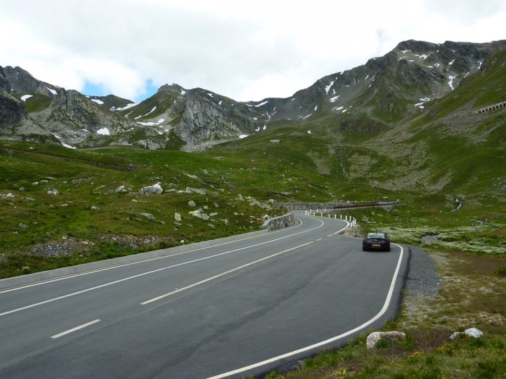 Trip of a Lifetime (picture heavy) - Page 1 - Aston Martin - PistonHeads - The image shows a winding paved highway cutting through a mountainous landscape. The mountains are rocky and mostly devoid of vegetation. A solitary car is traveling on the road, providing a sense of scale to the rugged terrain. The weather appears to be overcast with a gray sky. Along the right side of the highway, guardrails can be seen to ensure safety on the road.