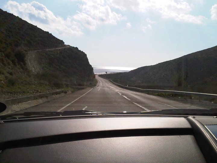 Hello from Cyprus! - Page 1 - Aston Martin - PistonHeads - This image captures a car's view of a curvy highway. The road appears to be empty and stretches out in front of the camera. To the right of the road, there is a tall rocky cliff, adding a sense of drama and vastness to the scene. The sky above is partially sunny, contributing a bright and inviting atmosphere to the image. There are also a few trees visible along the roadside, although they are not the primary focus of the image. The car's windshield is prominently displayed in the foreground, framing the road ahead and the landscape to the right.