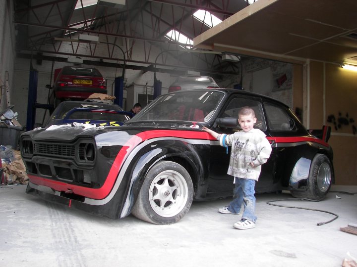 Pistonheads - In the image, a young boy is in the driver's seat of a black and red car parked indoors. He is leaning on the open door that displays a customized design. The car is parked next to a wall covered in white paper or a similar white material. There are other cars visible in the background, suggesting that this might be a workshop or a garage. Additionally, the presence of tools indicates a setting conducive to mechanical work or repairs.