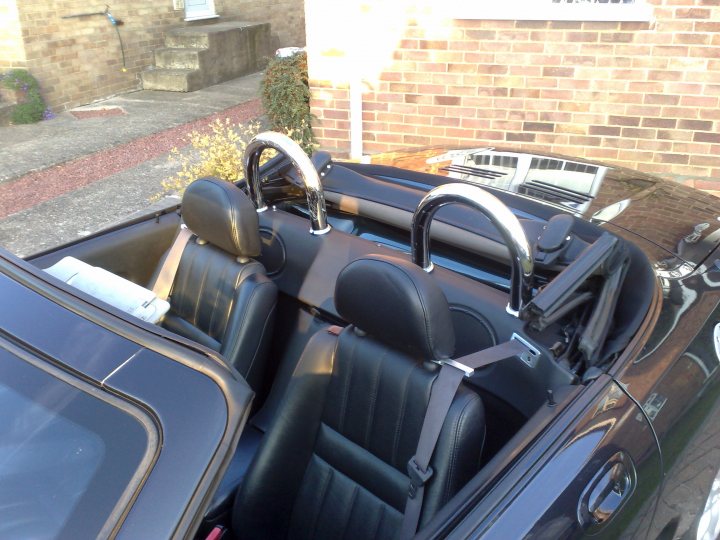 Pistonheads Hurt - The image shows the interior of a convertible. The car has a black top that is currently down. Inside, the vehicle features two front black seats that appear to have black seat belts. On the back of the front seats, there is a black leather material that appears to be part of a back seat that is not visible in this image. The sunroof has closed windows and the handle is in the upright position, hanging out of the rear window for convenience when opening. The car is parked outside, with its front end facing towards the camera.