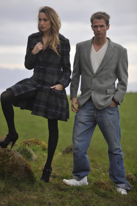 Horrid Pistonheads Chavs - In the image, a man and a woman are standing on a field covered with grass. The woman, positioned on the left, is wearing a long coat with a plaid pattern. She has her hand near the bottom of her coat, looking directly at the camera with a slight smile. On the right, the man is dressed in a patterned suit jacket over a white shirt. He has one hand in his pocket and is also looking at the camera with a neutral expression. The background reveals a hazy day with a clear sky above and hills in the distance.