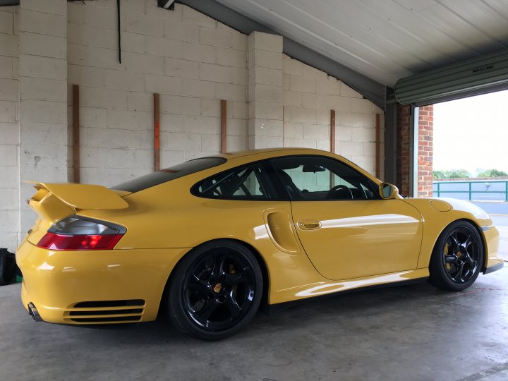 Black wheels - am I wrong? - Page 23 - General Gassing - PistonHeads - The image features a yellow sports car parked inside a building with concrete walls and a white ceiling. The car has a distinctive ducktail rear spoiler, glossy black rims, and is backed out of a garage, with the liftgate slightly open. The surrounding area suggests a classic or vintage setting, possibly a car show or a personal collection area, indicated by the presence of a stone wall, roll-up door, and exposed utility cables.