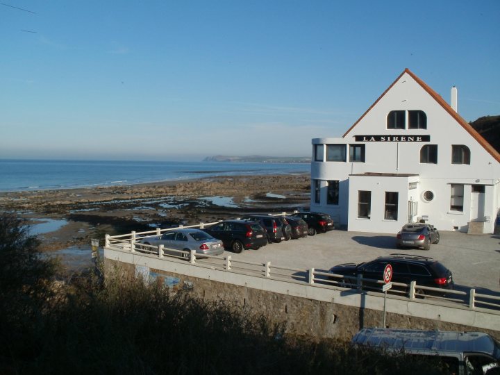 France Spotted Thread! - Page 1 - France - PistonHeads - The image is a daytime photograph depicting a white two-story building with a distinctive triangular roof situated on a promontory overlooking the ocean. In the foreground, there is a parking area with several cars parked alongside a concrete edge, providing a contrast to the building. A white fence runs parallel to the parking lot, separating the viewer's perspective from the property. The sky is clear, and the ocean stretches out to the horizon. The overall scene suggests a coastal town setting. There are no visible texts or markings in the image.