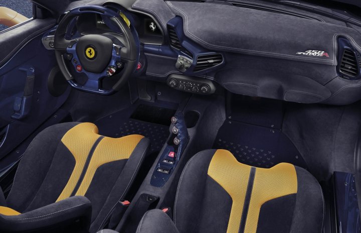 speciale spider is here  - Page 1 - Ferrari V8 - PistonHeads - The image is a photograph taken from the perspective of the driver's seat in a sports car. The vehicle interior is black, with accent colors of yellow and blue on the seats and the dashboard. The steering wheel is prominently equipped with buttons and has a dominant yellow Ferrari logo. The driver seat, visible in the foreground, has a blue back pad and a yellow lateral support pad. The car has a convertible top that is closed. The gear shift, lever, and gated manual transmission are also in view. The mood lighting of the interior illuminates the cockpit.